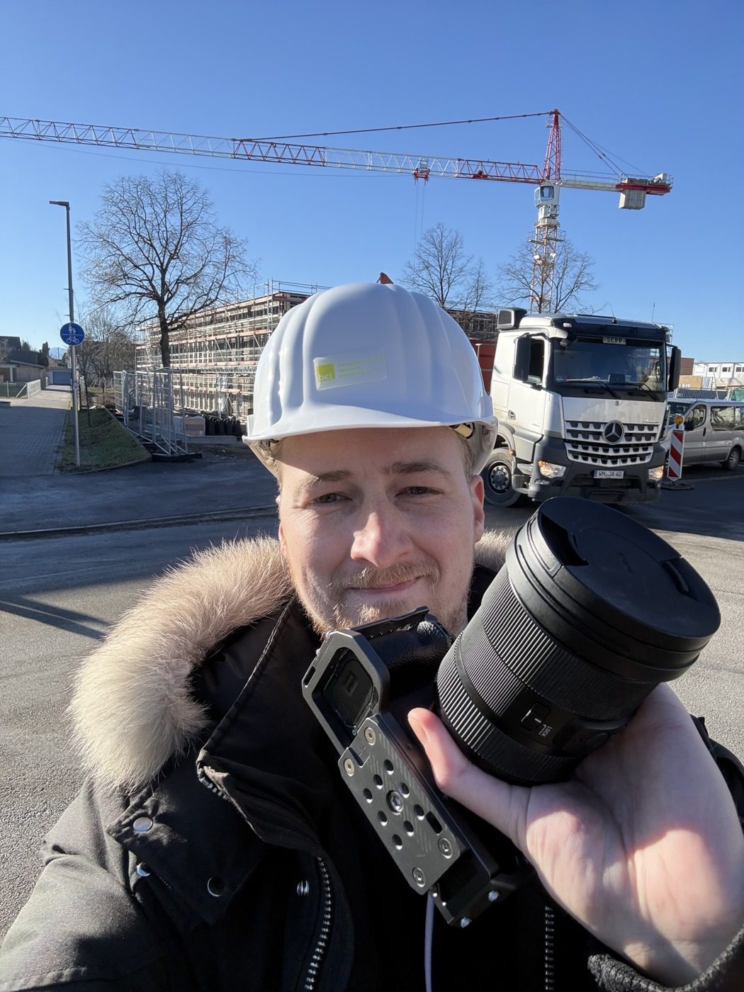 Mario Kaiser mit Bauhelm und Kamera auf einer Baustelle, dahinter Kran und Lastwagen